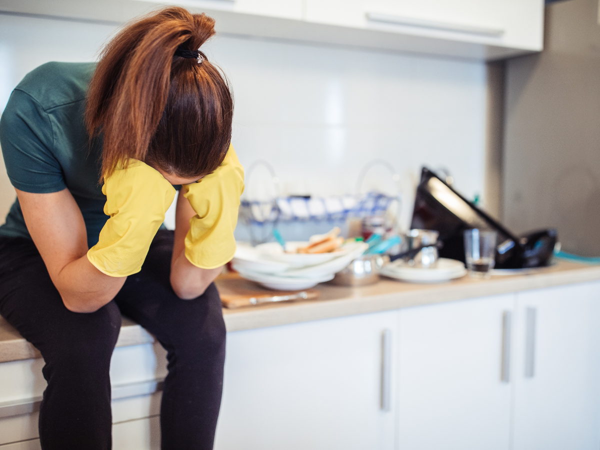 a woman sitting on her kitchen counter with dirty dishes overflowing in the sink. she has yellow gloves on and her head is down in her hands in frustration